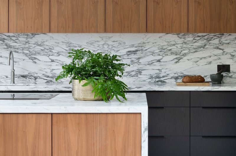Closeup at a Scandi kitchen design with dark wood base cabinets, lighter wood wall cabinets, veiny work surfaces, and a central island with a hob.