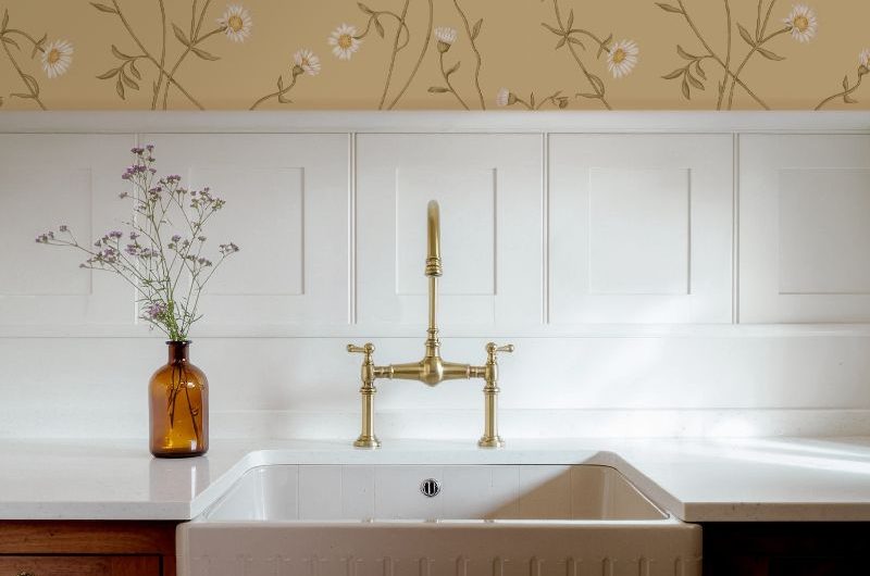 A sink zone with wood cabinets, a fluted sink, brass tap, wall panelling, and daisy patterned wallpaper.