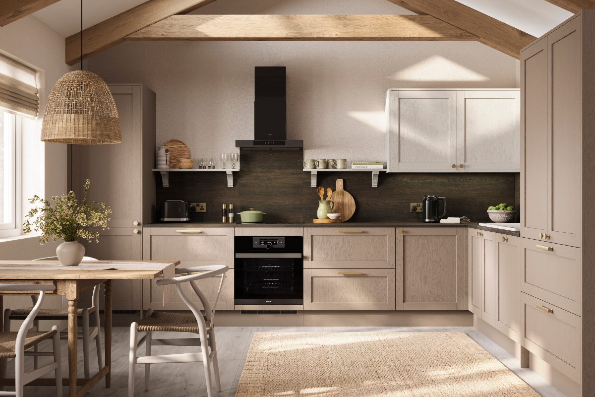 An L-shaped layout with classic Shaker cabinetry in neutral colours with a grainy finish, exposed beams, light wood flooring, and a dining zone near a window.