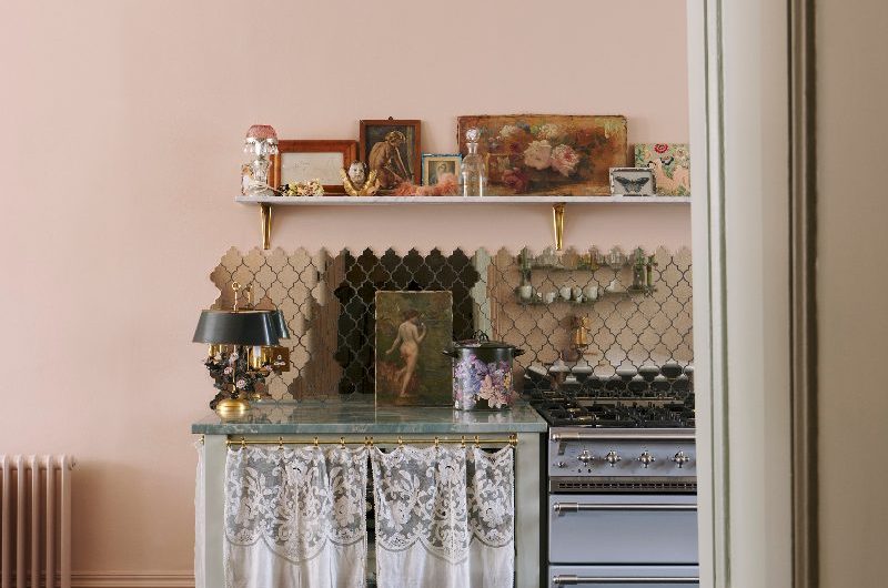 A classic scheme with mirrored tile splashback, a blue range cooker, open shelving, and storage concealed with curtains on brass rods, with pastel pink on the walls contrasted by dark wood flooring.
