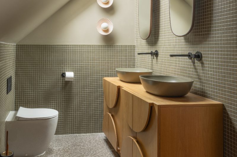 A minimalist scheme with a wood double vanity, with green countertop basins, mosaic wall tiles, and terrazzo flooring.