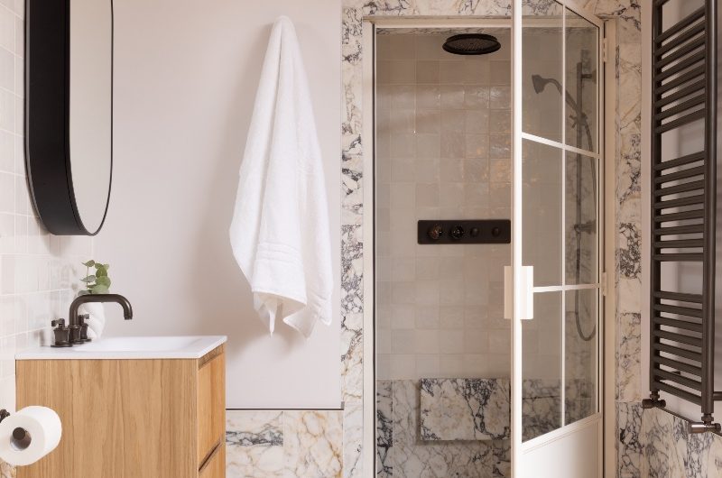 A luxurious bathroom with marble surfaces, a shower room with marble trims, and a vanity area with a wood unit, chunky tap, and mirror cabinet.