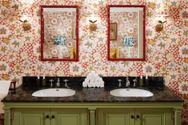 A double vanity unit painted in dark green with a black countertop, with floral wallpaper behind it and red bamboo-style mirror.