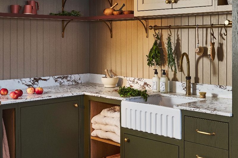 A green utility room with marble surfaces, neutral wall panelling, a butler sink with a brass tap, and a mix of open and closed wall-mounted storage.