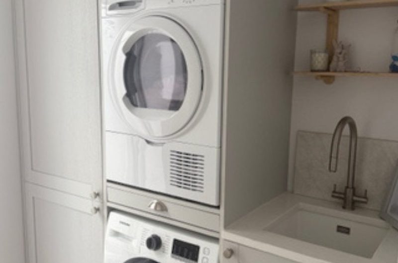 A utility room with stacked appliances, a sink and tao, neutral cabinetry, and open shelving.