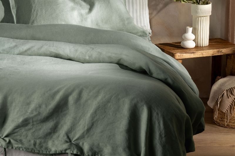 A serene sleep space with muted green bedding, limewash walls, and a wooden stool as a side table.