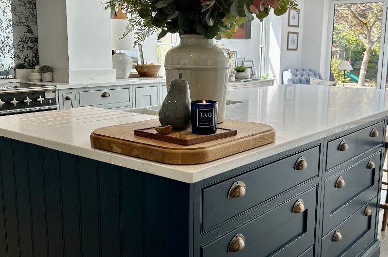 Closeup at an island with Shaker cabinets, brass handles, white worktops, and layered lighting.