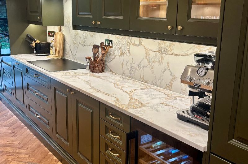 A dark green, classic kitchen renovation with built-in appliances, lighting and marbled surfaces.