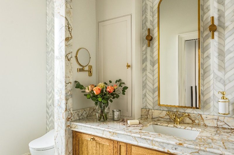 A bespoke oak vanity with warm marble, leaf-like marble wall tiles, inset sinks with gold tap, and patterned flooring looking into the toilet area through a curved entryway.