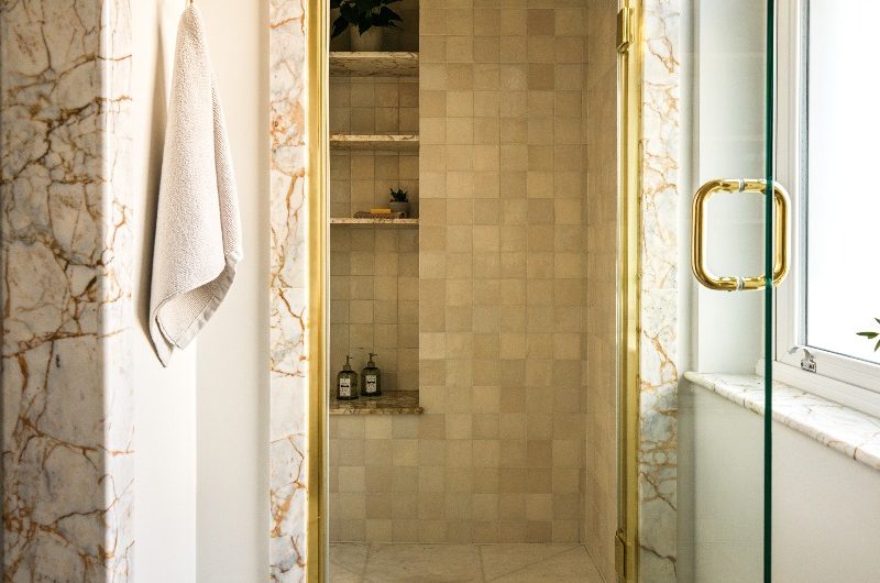 A luxury ensuite with zellige-like tiles, open shelving, a glass door, and gold fittings.
