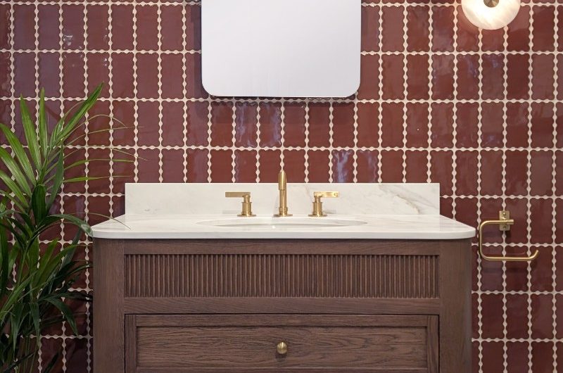 A boutique bathroom design with a wood vanity and a mix of tiles in oxblood and white, and gold fittings and fixtures.