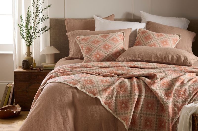 A spring bedroom with pale terracotta linen, patterned cushions and throw, wood beside table, and rattan rug.