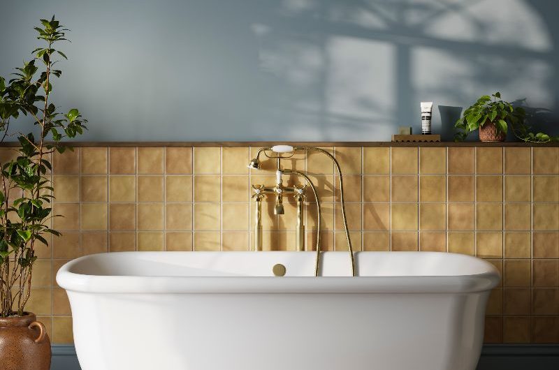 A freestanding tub with a glossy finish in a modern, colourful scheme, with yellow zellige tiles on the back wall, blue paint, and mosaic flooring.