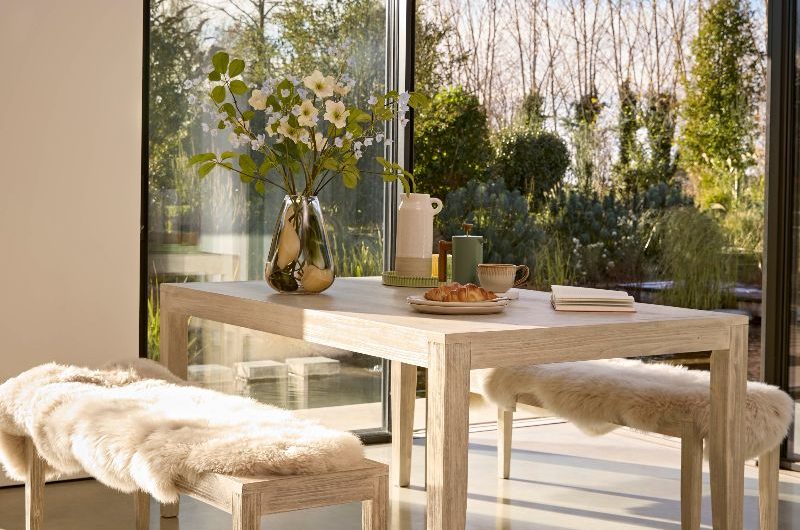 A dining room with wide expanse of glazing, with a limewash dining table, benches, and faux fur seating covers.