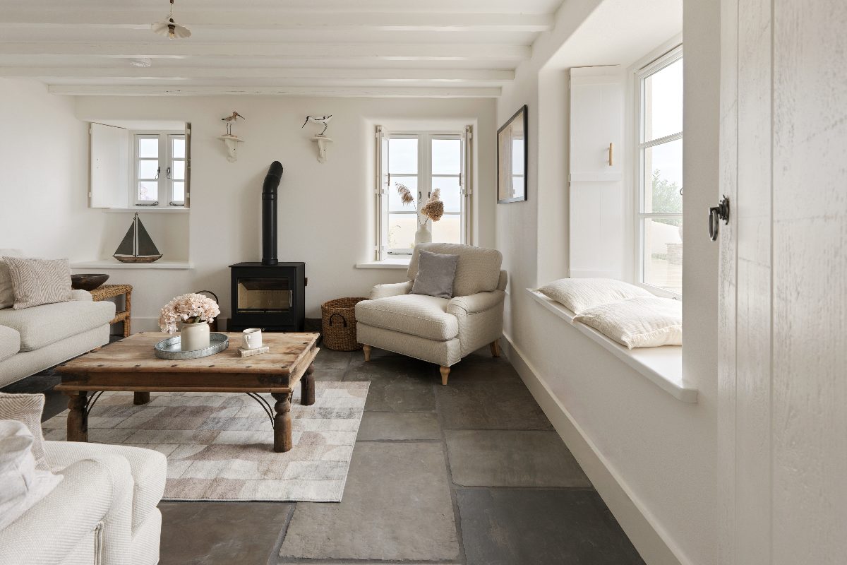 A coastal living room with stone flooring, whitewashed alls, neutral furnishings, a log burner, and open windows.