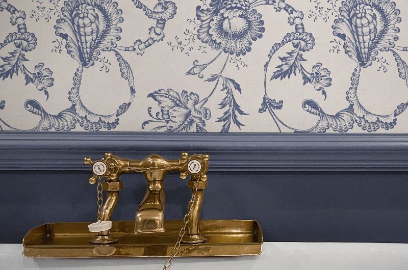 Closeup at a bathroom with blue panelling, patterned wallpaper, and a tub with brass filler.