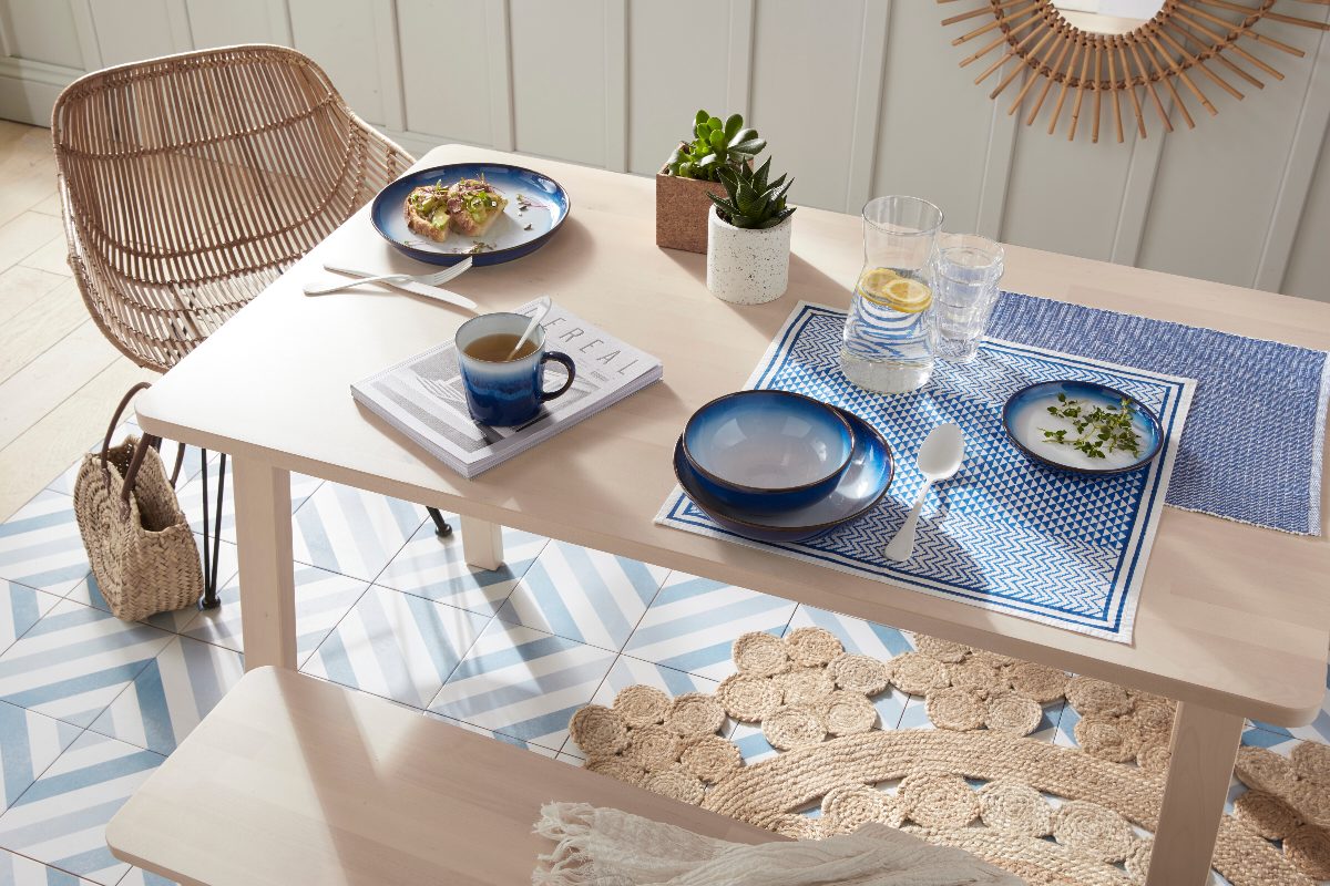 A coastal dining room with a light oak table and bench, blue and white striped floor tiles, ombre blue crockery and a rattan rug.