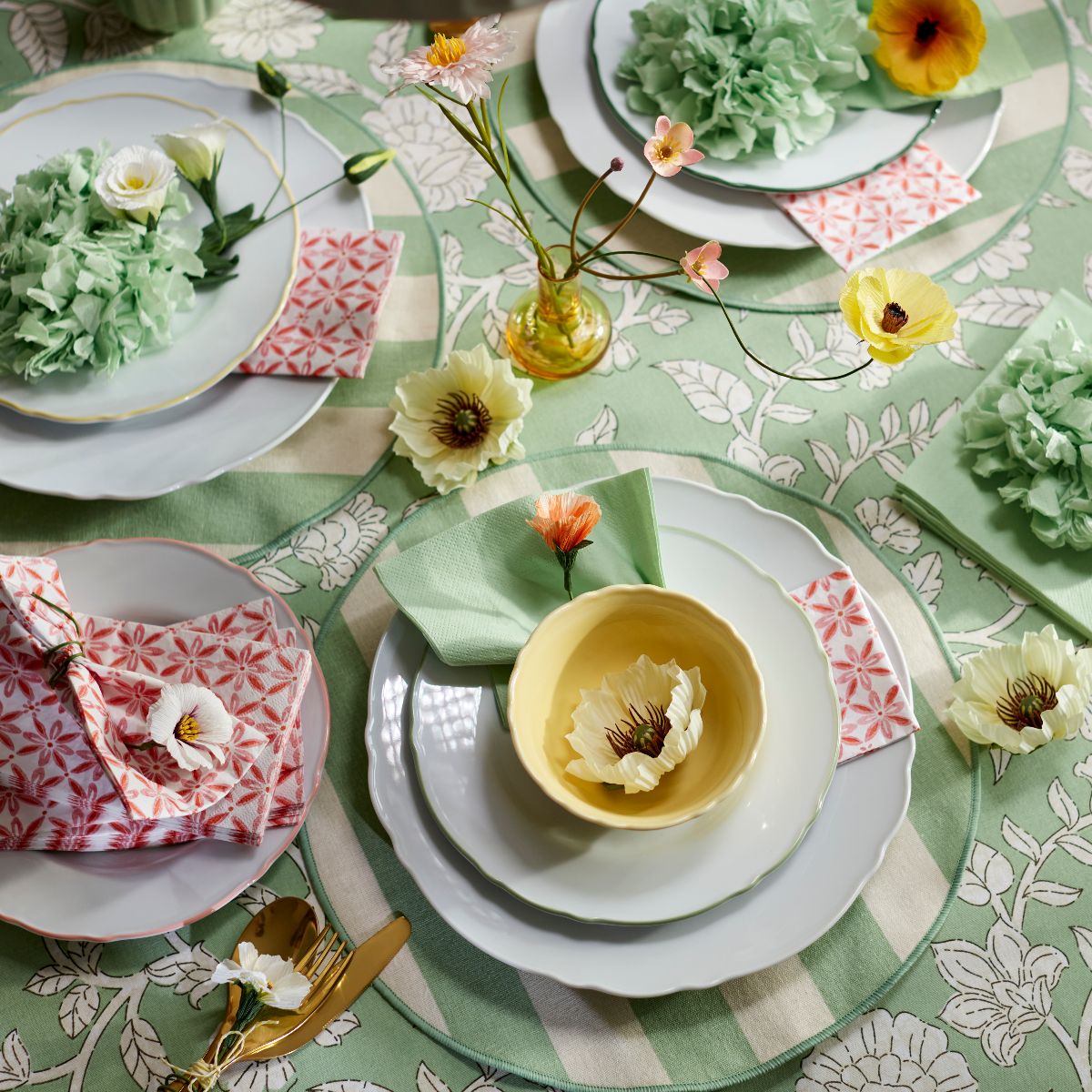 An Easter tablescape with green floral tablecloth, striped placemats, white ceramics, and gold cutlery.