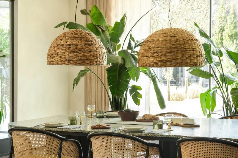 A zen dining room with house plants, a dark table, rattan chairs, and giant dome pendants above it.