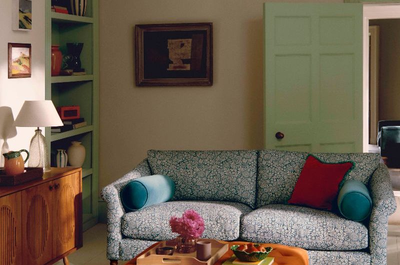 A heritage-inspired living room with a floral sofa, an orange footstool, wood sideboard, and green bookcase.