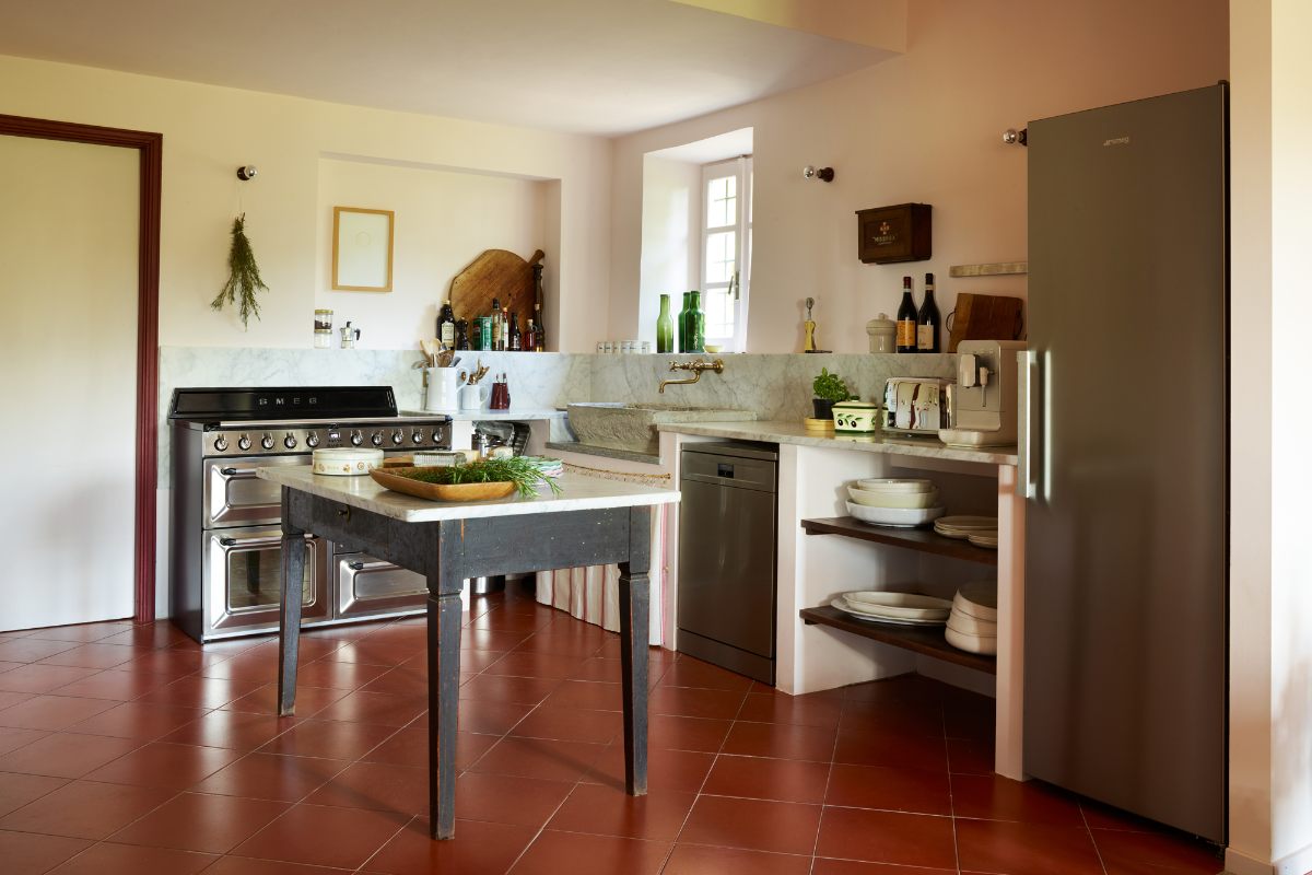 A kitchen design in Italy with Smeg appliances, dark red flooring, a stone sink, and marbled surfaces.
