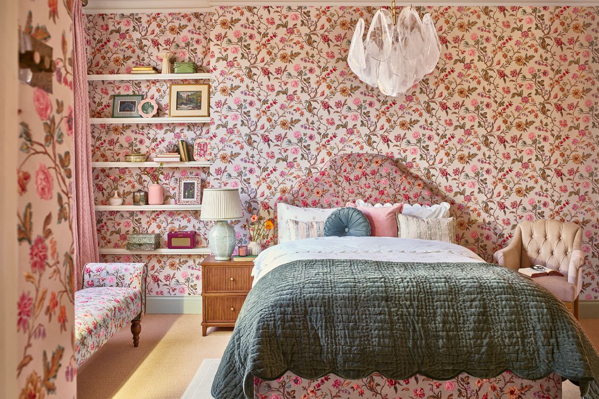 A romantic bedroom with floral wallpaper, a matching bed frame and ottoman, with open shelving, and scalloped bedding and throw.