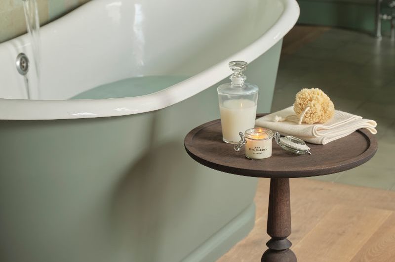 A calming bathroom with green zellige tiles, a green painted freestanding tub, a dark wooden side table, and wood flooring.
