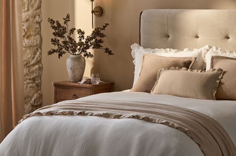 A calm, sophisticated bedroom with exposed stone on the wall, a cream upholstered bed, and white tactile linen.