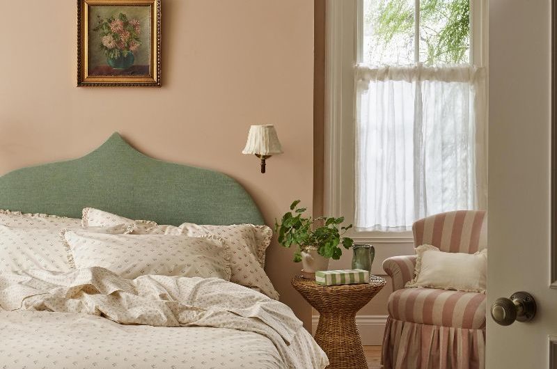 A spring summer bedroom design with a green headboard, floral bedding, a rattan side table, striped armchair, pale pink wall paint, and a white airy cafe curtain on the window.