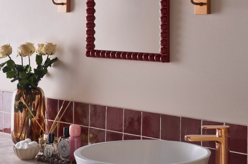 Closeup at a terrazzo countertop with a white basin, copper tap, and a pink splashback and matching bobbin mirror.