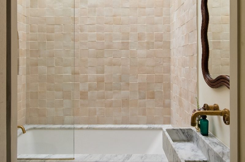 A serene bathroom with zellige wall tiles, a fitted bath with marble surround, and a marble basin with a rattan mirror above it.