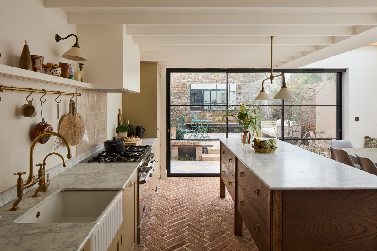 View of the new kitchen with steel glazing, terracotta flooring, a freestanding island topped with marble, and a run of units on the left side.