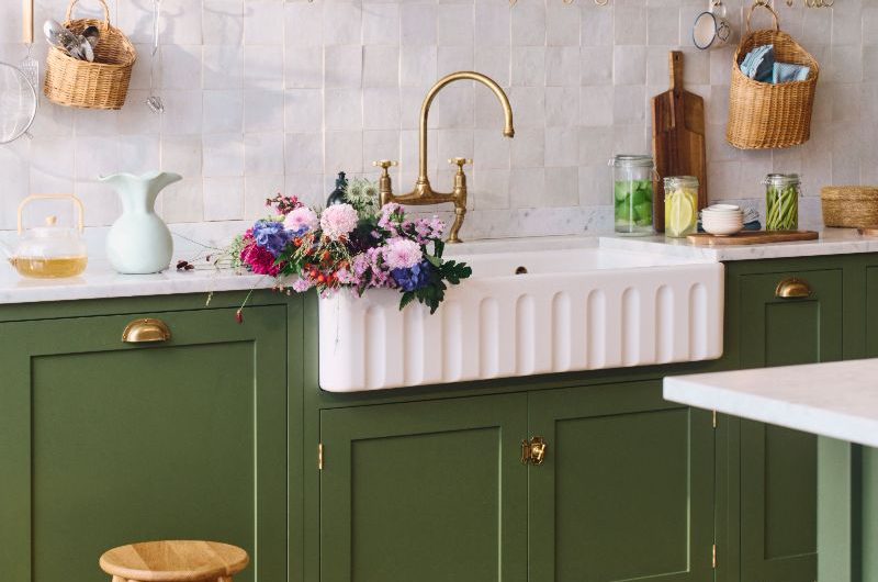 A dark green kitchen with Shaker cabinets, wood flooring, a fluted butler sink, brass tap, zellige tiles, and open shelving.