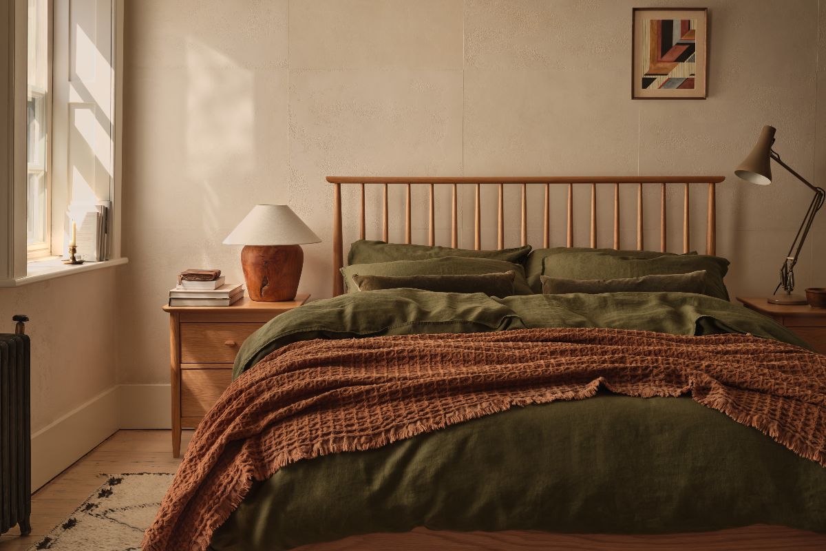 A calm bedroom with olive green bed linen, a waffle throw in a terracotta colour and textures walls in the background.
