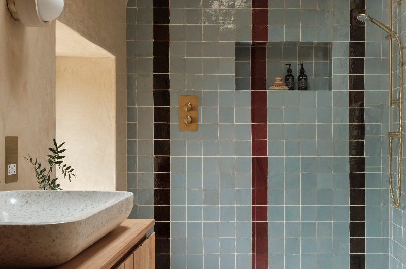 A modern, tactile bathroom with a colourful shower, plaster on the walls, a wood vanity, and a speckled basin and flooring.