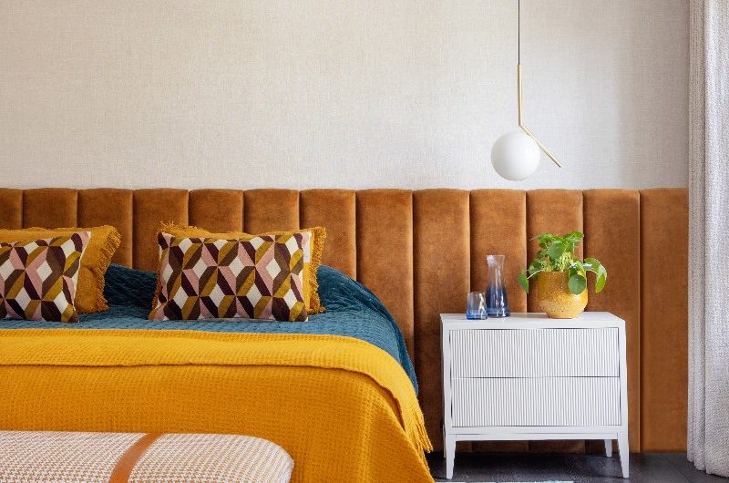 A colourful bedroom design with an extra large headboard, coloured rug, fluted side table, upholstered footstool, and pendant lighting.