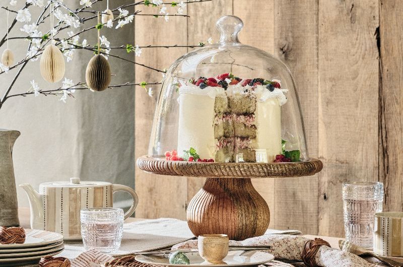 A rustic Easter tablescape with mango wood decor, patterned ceramics, and a wooden cake stand with a tall dome.