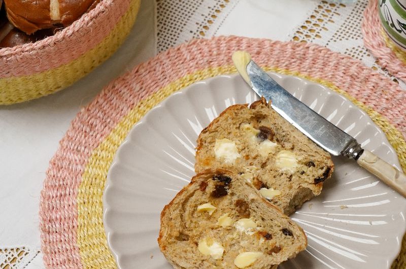 A breakfast table with pink and yellow circular placemats, a butter dish, and hot cross buns.