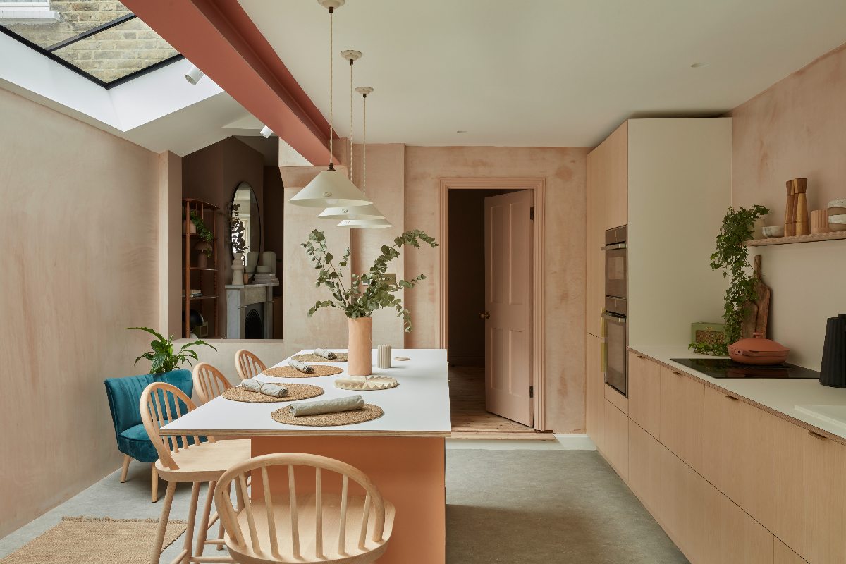 A family kitchen design with a run of cabinets on the right and a central island with seating and pendant lights above it, with warm plaster on the walls and a glazed roof.