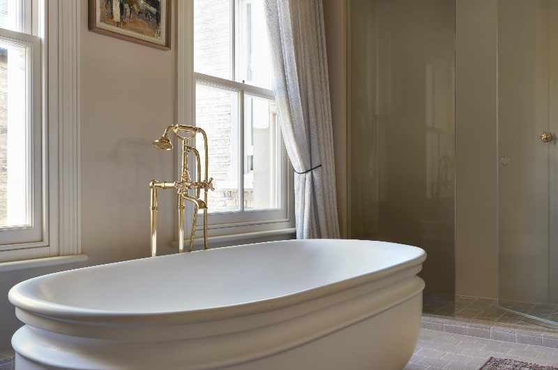 A freestanding tub with curves, a gold bath filler, and sash windows.