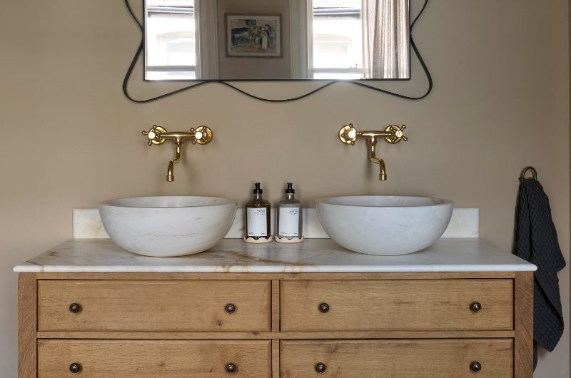 A double vanity made from wood, with a veiny worktop, countertop basins, gold taps, and a statement mirror.