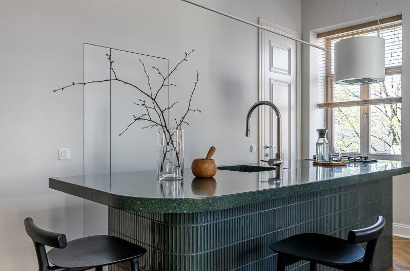 Closeup at an island with green cement tiles, a green worktop, black stools, and a sink and tap.