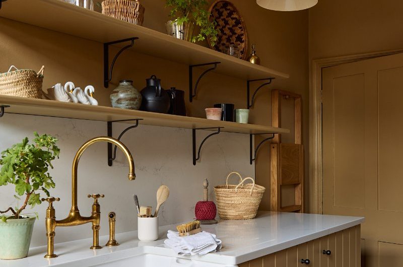 A colour drenched kitchen design in a warm mustard yellow, with a sink zone with a butler sink and brass tap, with open shelving and black fittings.