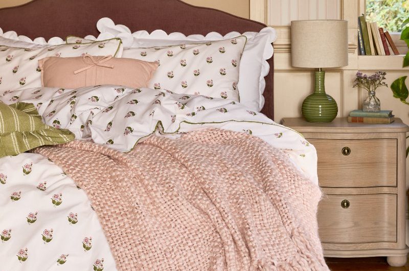 A rustic bedroom with wood flooring, bedside table, upholstered bed with white and floral linen, and striped wallpaper.