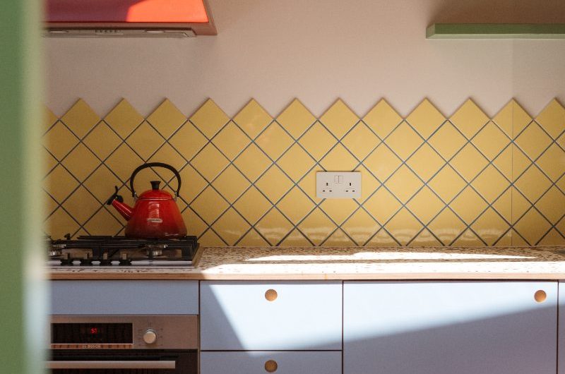 A modern scheme with yellow splashback tiles, terrazzo worktop, sky coloured cabinets, and a red extractor canopy.