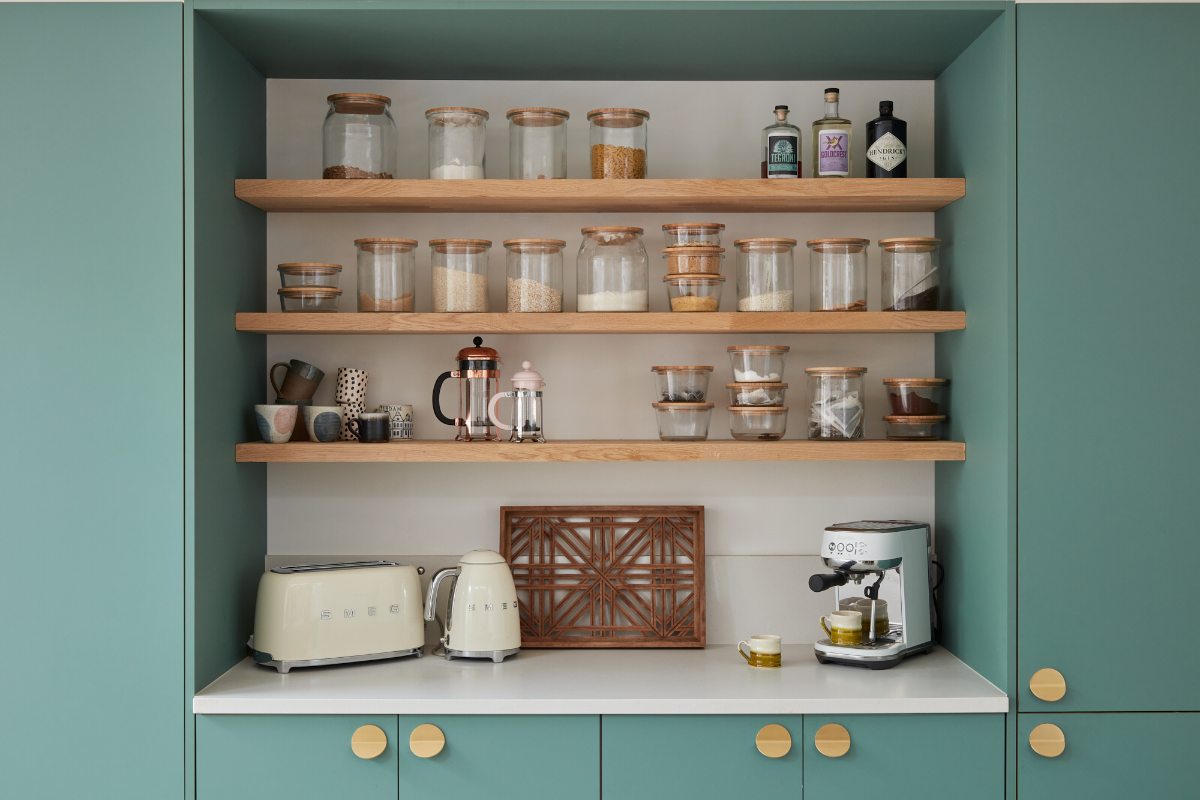 A coffee station with green units, oak shelves, and neutral worktops.