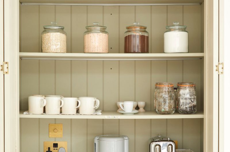A coffee station with a sage green interiors, open shovelling, white cups, and metallic appliances.
