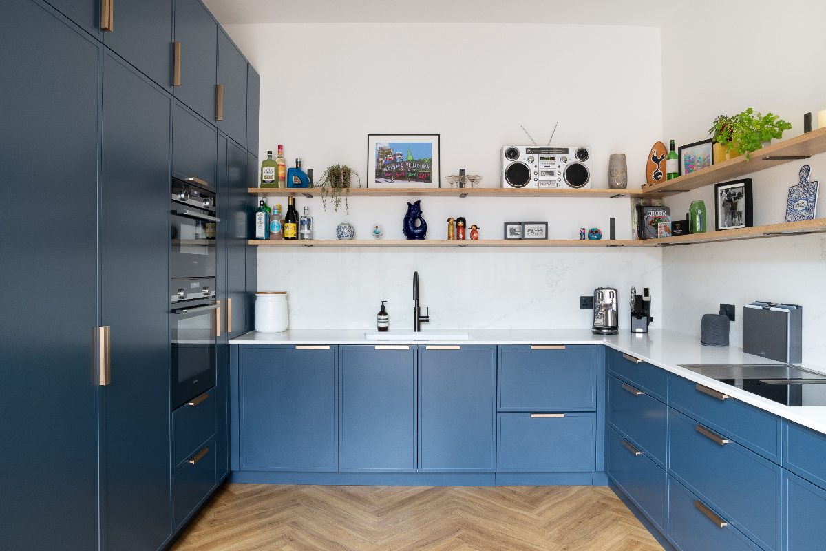 A U-shaped kitchen with blue units, metallic handles, oak open shelving, and wood flooring.