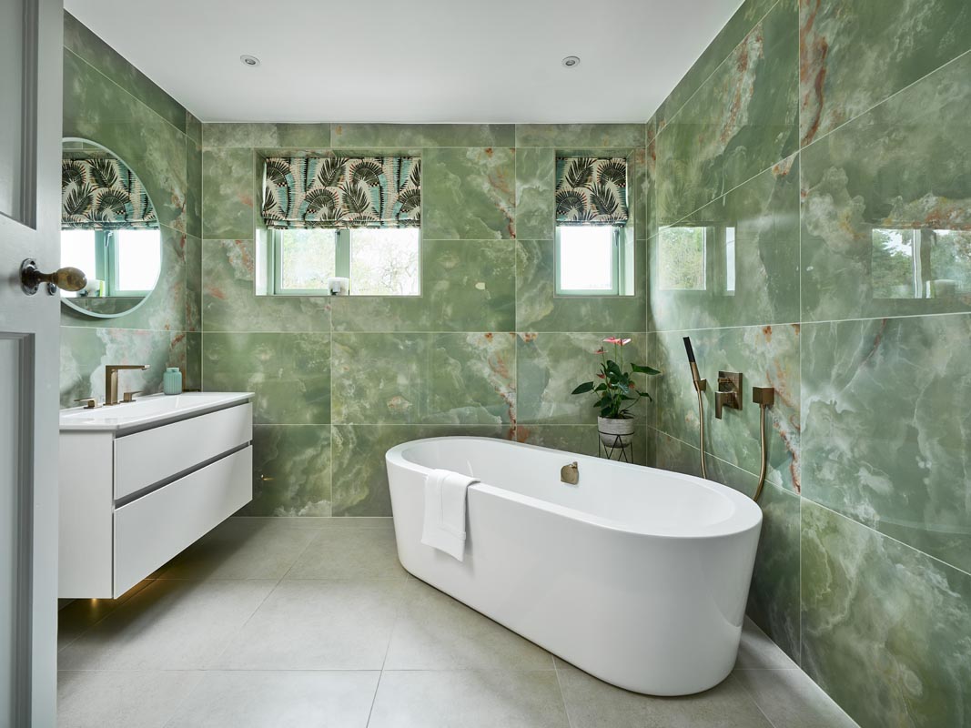 Modern ensuite design with floor-to-ceiling green onyx-effect tiles, a freestanding tub, a vanity area and gold brassware.