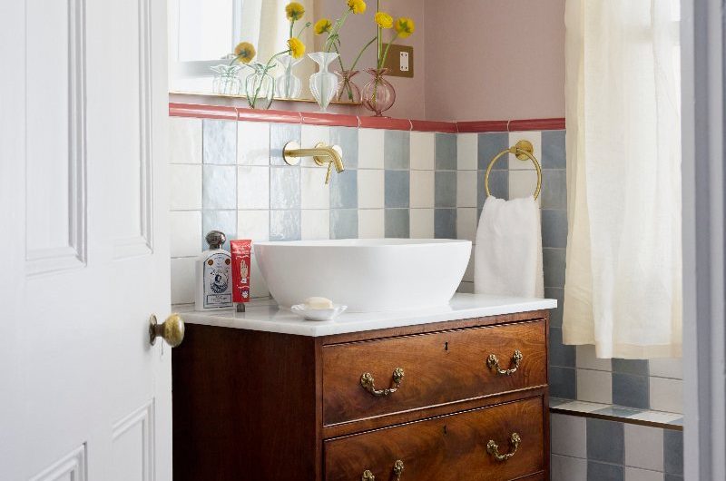 A colourful bathroom with striped tiles, pale pink walls, vintage vanity unit, white countertop basin and gold accents.